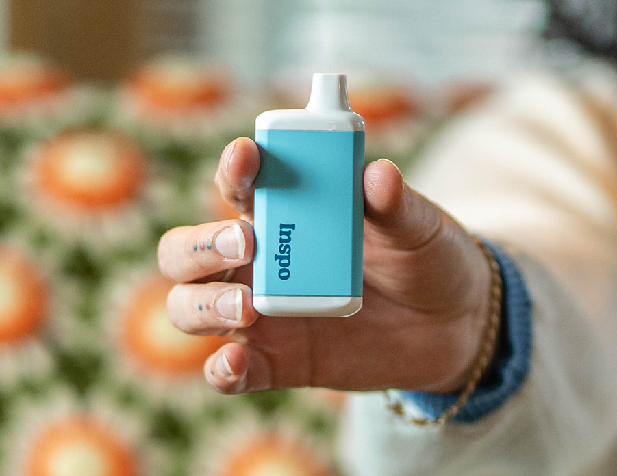 A hand holds up a light blue Inspo vape device, showcasing its sleek and compact design. The background features a blurred, retro-style floral pattern with orange and white flowers. The close-up shot emphasizes the device's portability and stylish aesthetic.