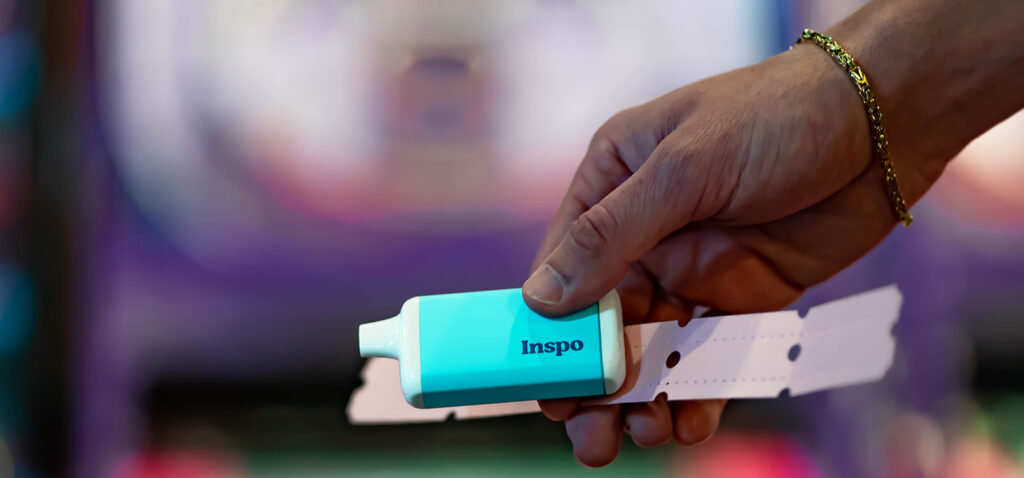 A close-up of a hand holding a light blue Inspo vape device alongside a strip of arcade tickets. The blurred background features colorful lights, evoking a fun, nostalgic gaming atmosphere. The image connects the Inspo brand with a playful, rewarding experience.