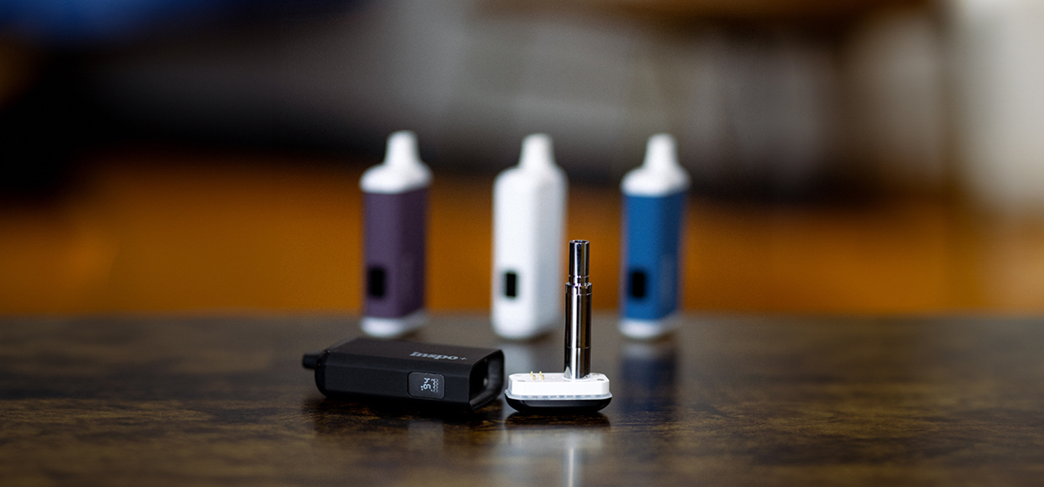 A close-up of a black Randy's Inspo vaporizer battery with a detachable cartridge attachment, placed on a wooden surface. In the softly blurred background, three other Inspo vaporizers in purple, white, and blue stand upright. The image highlights the sleek design and variety of color options in the Inspo vaporizer line.