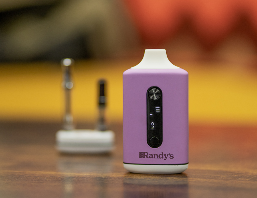 A lavender-colored Randy's Inspo Dual vaporizer stands upright on a wooden surface, its digital display illuminated. In the blurred background, two vape cartridges rest on the table, emphasizing the device’s compatibility. The sleek design and soft color make the vaporizer visually appealing, blending technology with style.