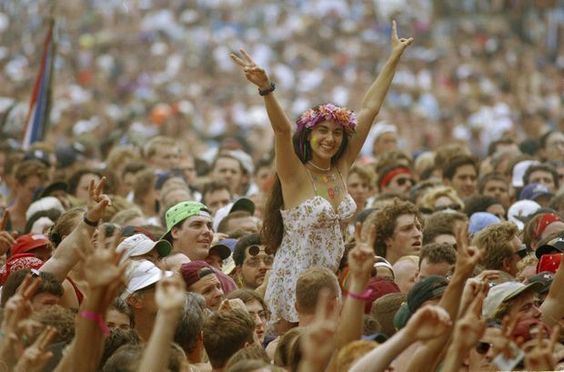 Smiling woman at a crowded outdoor festival raising her hands in peace signs, surrounded by a lively crowd.