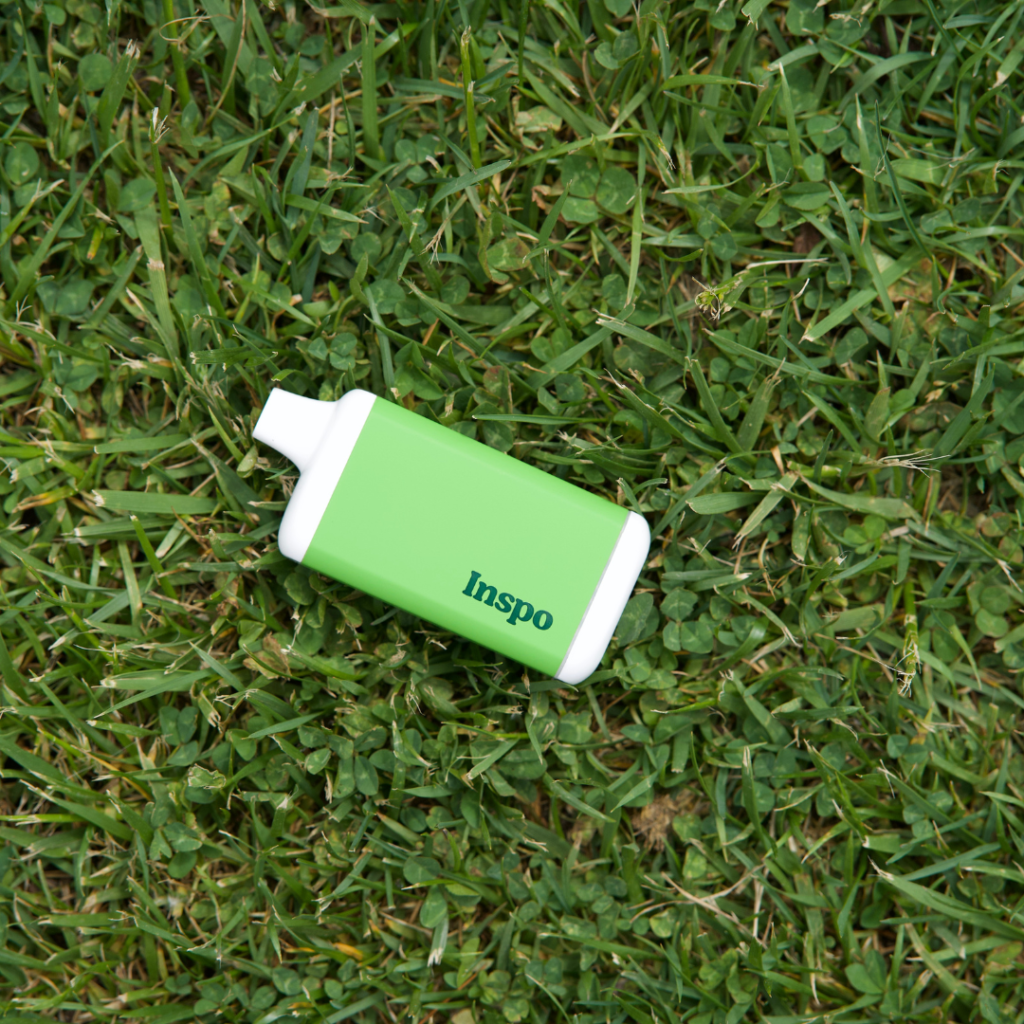 A green Inspo vape device rests on a bed of fresh grass, blending seamlessly with the natural background. The sleek, compact design of the vape stands out with its white accents and bold branding. The image conveys a connection to nature, possibly hinting at eco-conscious vaping choices vs disposable vapes